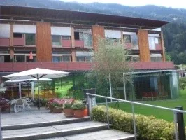 Ein modernes Gebäude mit Glasfenstern und einem roten Vordach, umgeben von Grün und einem Berg im Hintergrund.