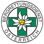 Eine weiße Blume mit gelben Blütenblättern ist auf einem grünen Kreuz platziert, das von einem weißen Kreis umgeben ist. Die Worte 'Bergrettungsdienst Osterreich' sind in einer gebogenen Weise um den Kreis herumgeschrieben.
