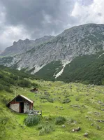Eine kleine Holzhütte steht auf einem grasigen Hang mit einem Gebirgszug im Hintergrund.