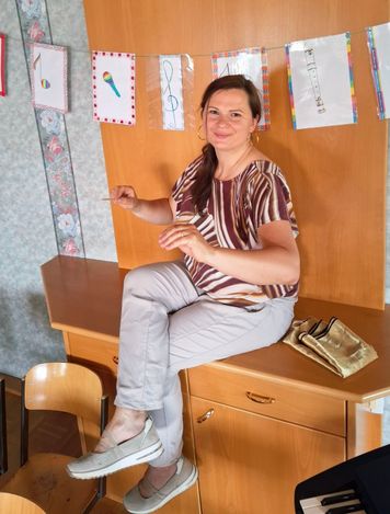 Eine Frau in einem gestreiften Shirt und grauer Hose sitzt auf einem Holzschreibtisch, hält einen Stock und lächelt für ein Foto. Hinter ihr ist eine Holzwand mit bunten Zeichnungen und Papieren zu sehen.