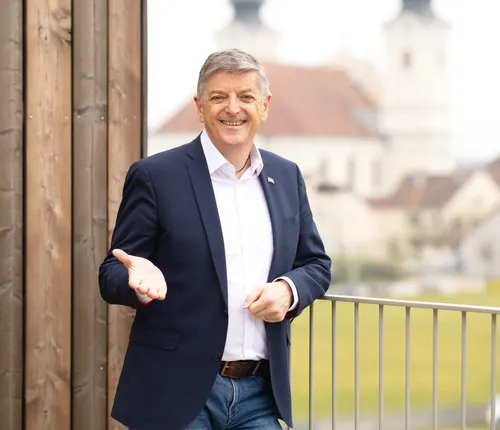 Ein Mann in einem dunkelblauen Anzug steht draußen, gestikuliert mit der Hand und lächelt. Er steht auf einem Balkon mit einer Brüstung. Hinter ihm befindet sich eine Holzwand. Im Hintergrund steht ein Gebäude und ein grünes Feld.