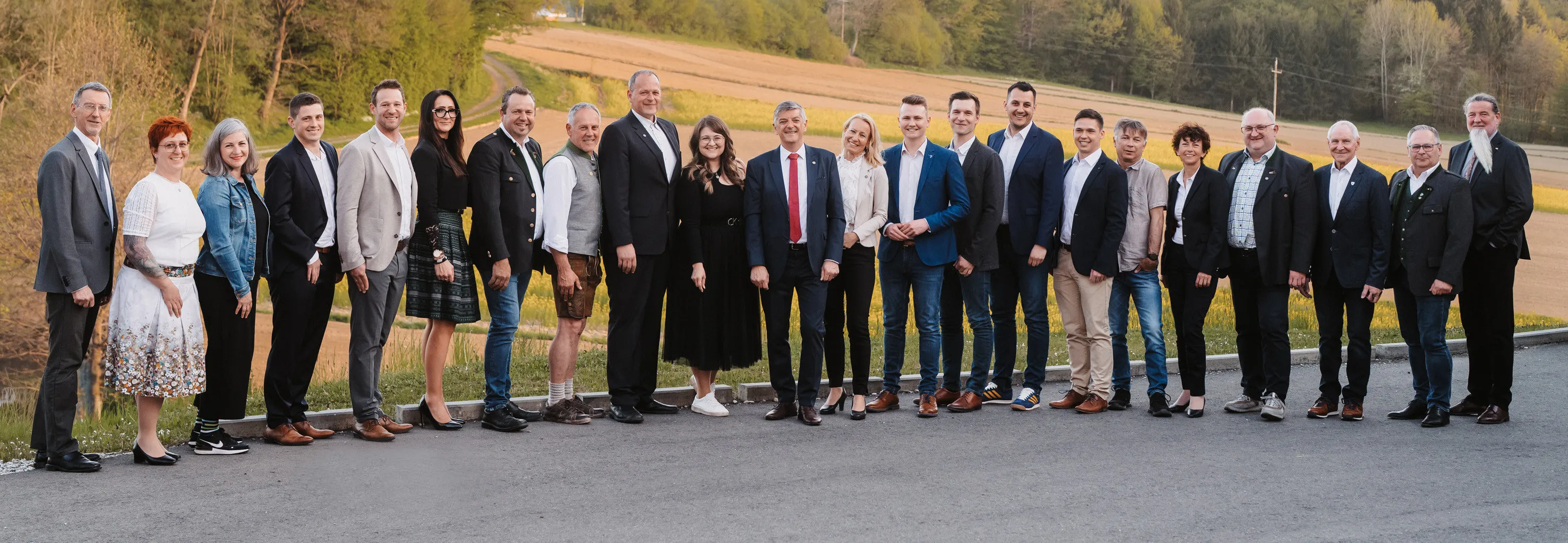 Eine Gruppe von Menschen in formeller Kleidung steht auf einer asphaltierten Straße, lächelt und posiert für ein Foto. Sie stehen auf einer Betonplattform mit einem Grasfeld und Bäumen im Hintergrund.