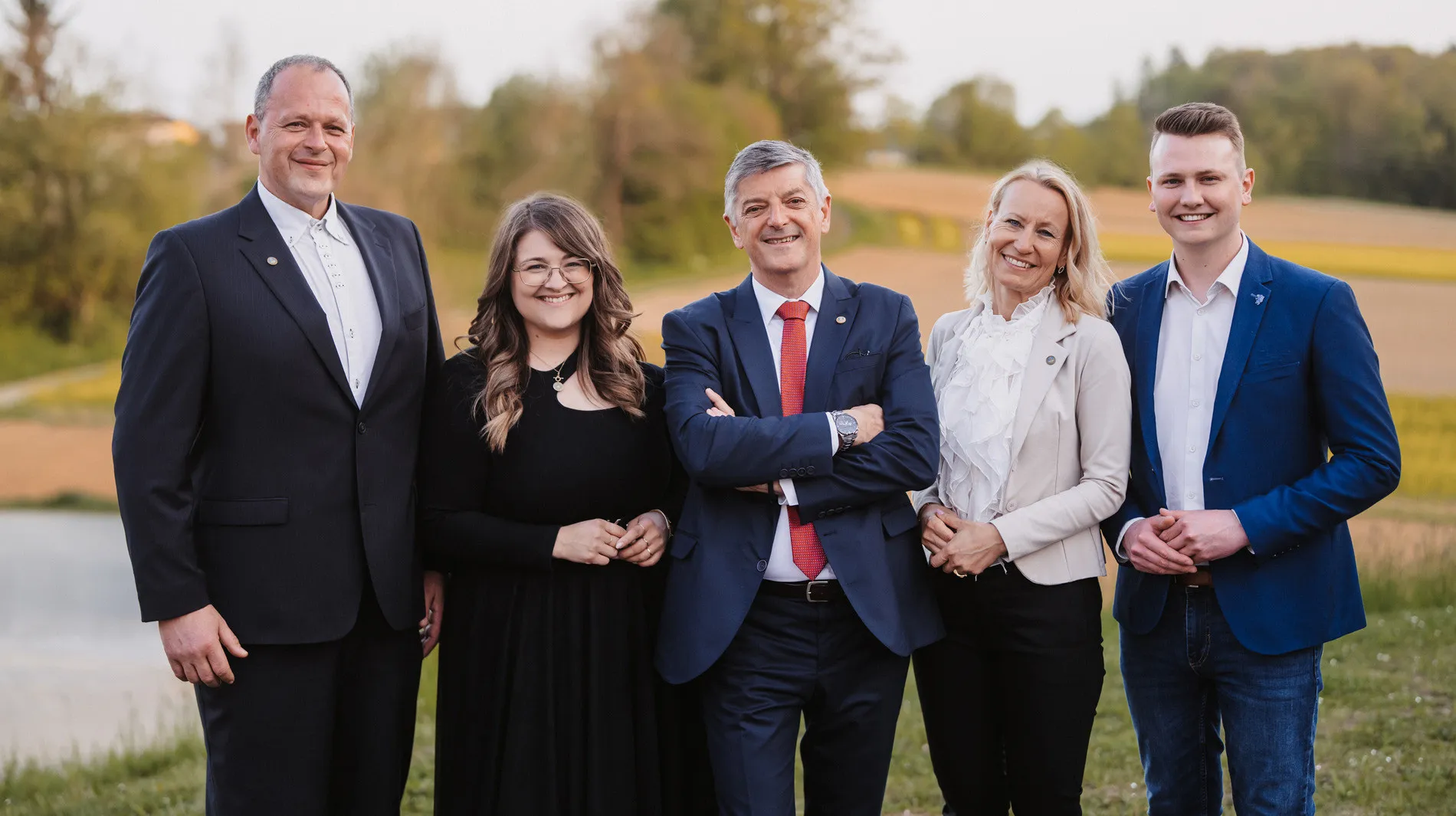 Fünf Personen stehen auf einem Feld. Zwei Frauen und drei Männer. Der Mann in der Mitte lächelt und hat die Arme verschränkt.