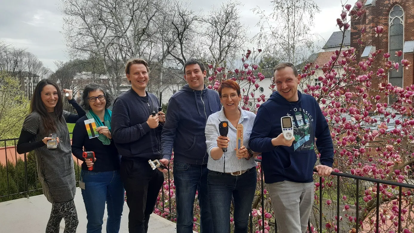 Eine Gruppe von sechs Personen, in Freizeitkleidung gekleidet, lächelt und posiert für ein Foto vor blühenden Bäumen. Jede Person hält verschiedene elektronische Geräte in den Händen.