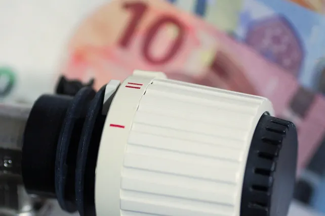 Eine Nahaufnahme eines weißen Thermostats mit einer roten Linie darauf. Ein verschwommenes Bild einer Zehn-Euro-Banknote ist im Hintergrund.