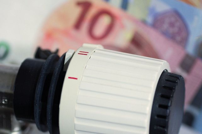 Eine Nahaufnahme eines weißen Thermostats mit einer roten Linie darauf. Ein verschwommenes Bild einer Zehn-Euro-Banknote ist im Hintergrund.