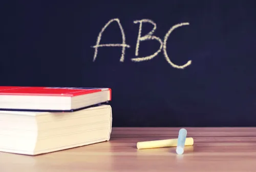 Eine Tafel zeigt das Wort ABC in Kreide geschrieben, mit zwei Büchern auf einem Holztisch und einem gelben Kreidestück in der Nähe.