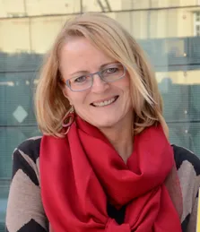 Eine Frau mit kurzen blonden Haaren und Brille, lächelnd und mit einem roten Schal, steht vor einer verschwommenen Glaswand.