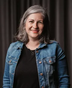 Eine lächelnde Frau mit grauen Haaren trägt ein schwarzes Shirt und eine Jeansjacke mit braunen Knöpfen. Sie steht vor einem dunklen Hintergrund.