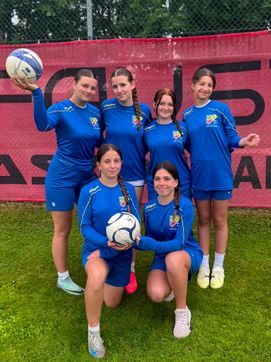 Fünf weibliche Fußballerinnen in blauen Trikots posieren für ein Foto. Eine Spielerin hält einen Fußball, während die anderen lächeln. Sie stehen auf einem Rasenfeld mit einem roten Zaun im Hintergrund.