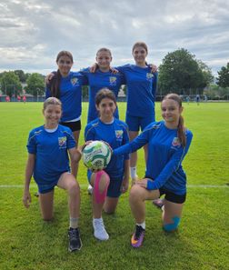 Fünf junge Mädchen in passenden blauen Trikots posieren für ein Foto auf einem Grasfeld. Sie halten einen Fußball und lächeln. Im Hintergrund sind weitere Menschen und Bäume zu sehen.