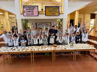 Eine Gruppe von Schülern posiert für ein Foto vor einem langen Tisch voller Essen. Sie heben ihre Hände in die Luft. Der Tisch hat viele Teller mit verschiedenen Speisen. Es gibt auch Tassen und Flaschen auf dem Tisch. Dahinter hängen zwei Banner auf dem Balkon.