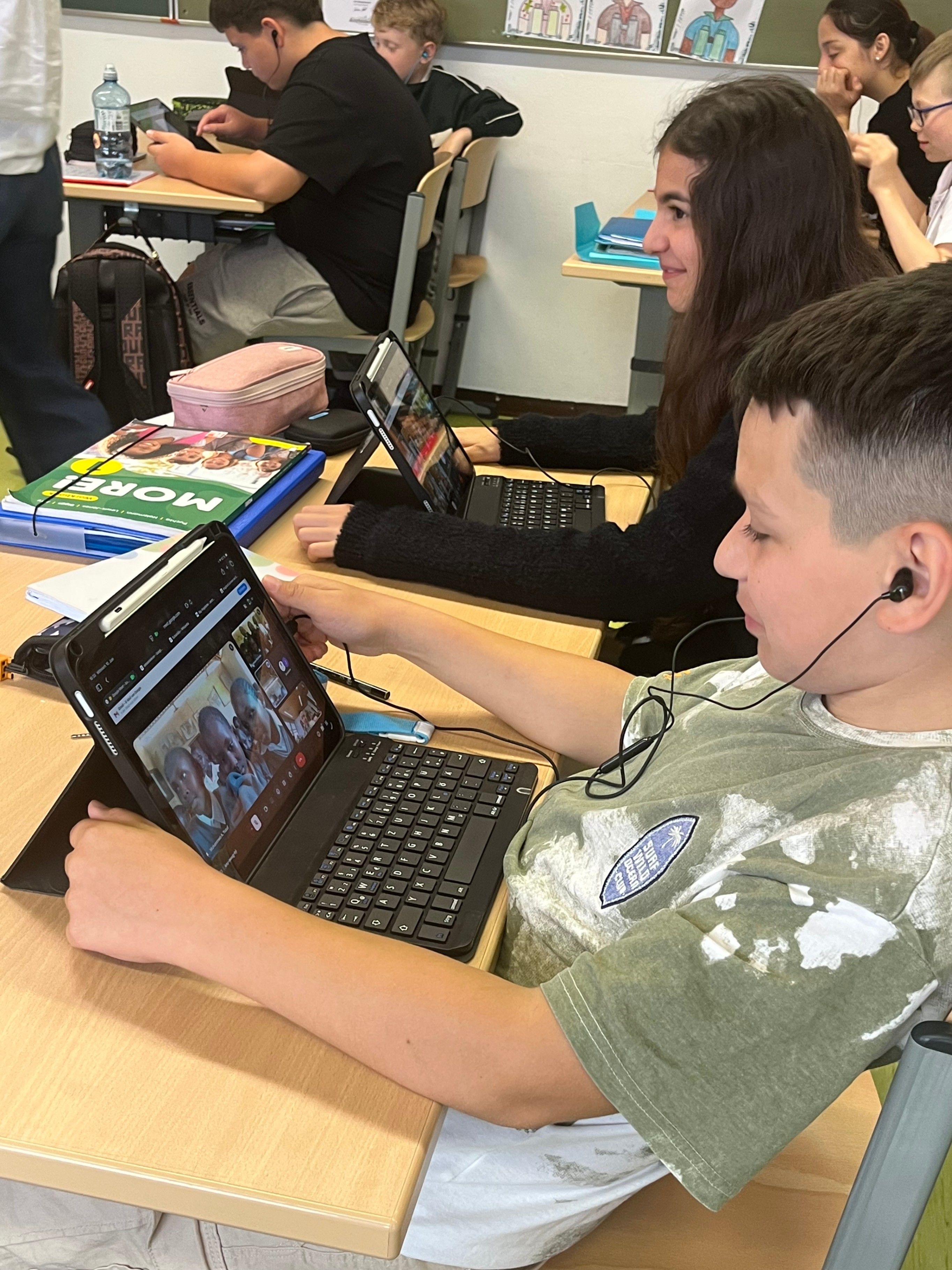 Ein Junge arbeitet in einem Klassenzimmer an einem Tablet. Er hat Kopfhörer auf. Hinter ihm arbeitet ein Mädchen an einem Laptop. Andere Schüler sind anwesend.