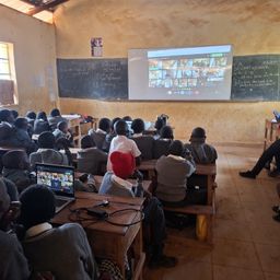 Schüler in einem Klassenzimmer nutzen Laptops für eine Videokonferenz, wobei ein Beamerbildschirm die Sitzung anzeigt. Hinter ihnen stehen Tafeln mit Notizen.