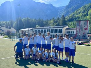 Ein Sportteam posiert für ein Foto auf einem Feld mit Bergen im Hintergrund. Sie tragen passende weiße T-Shirts und blaue Shorts.