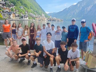 Eine Gruppe junger Menschen posiert für ein Foto auf einem Steg mit Bergen und einem See im Hintergrund. Einige tragen Sonnenbrillen und Turnschuhe, und einer trägt ein Emirates-Shirt.