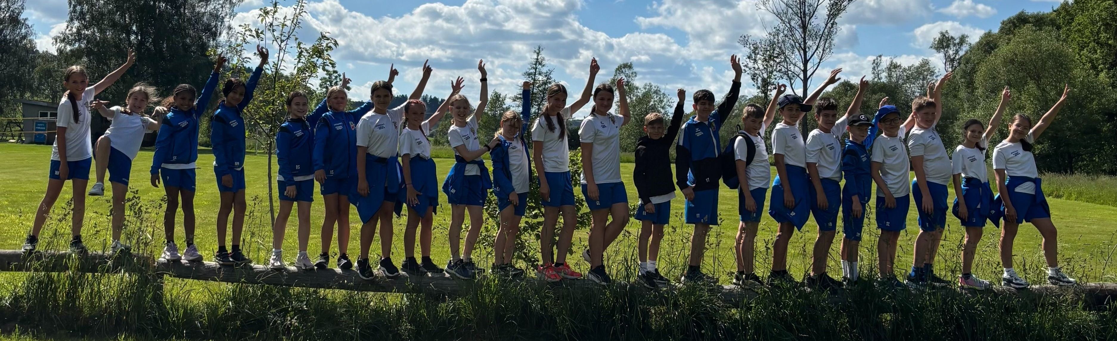 Eine Gruppe junger Schüler in identischen Uniformen steht auf einem Grasfeld. Sie alle lächeln und heben ihre Hände. Sie stehen auf einem Holzbrett in der Nähe einiger Pflanzen.
