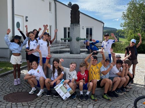 Eine Gruppe von Kindern posiert für ein Foto vor einem Brunnen, mit einem Hintergrund aus einem weißen Gebäude und Bergen.