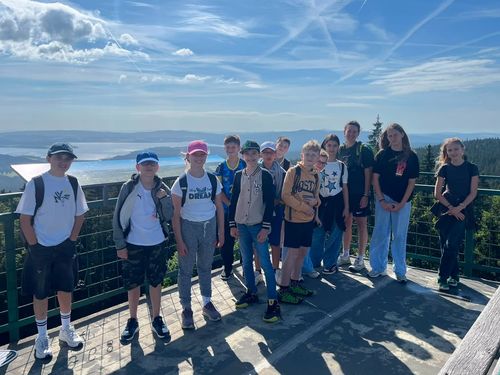Eine Gruppe junger Menschen steht zusammen auf einem Balkon und lächelt für ein Foto. Sie tragen Freizeitkleidung und stehen vor einer wunderschönen Landschaft.