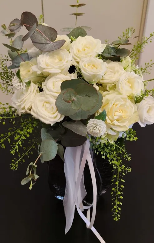 Ein Strauß weißer Rosen mit grünen Blättern und einer weißen Schleife, arrangiert in einer schwarzen Vase auf einer dunklen Oberfläche.