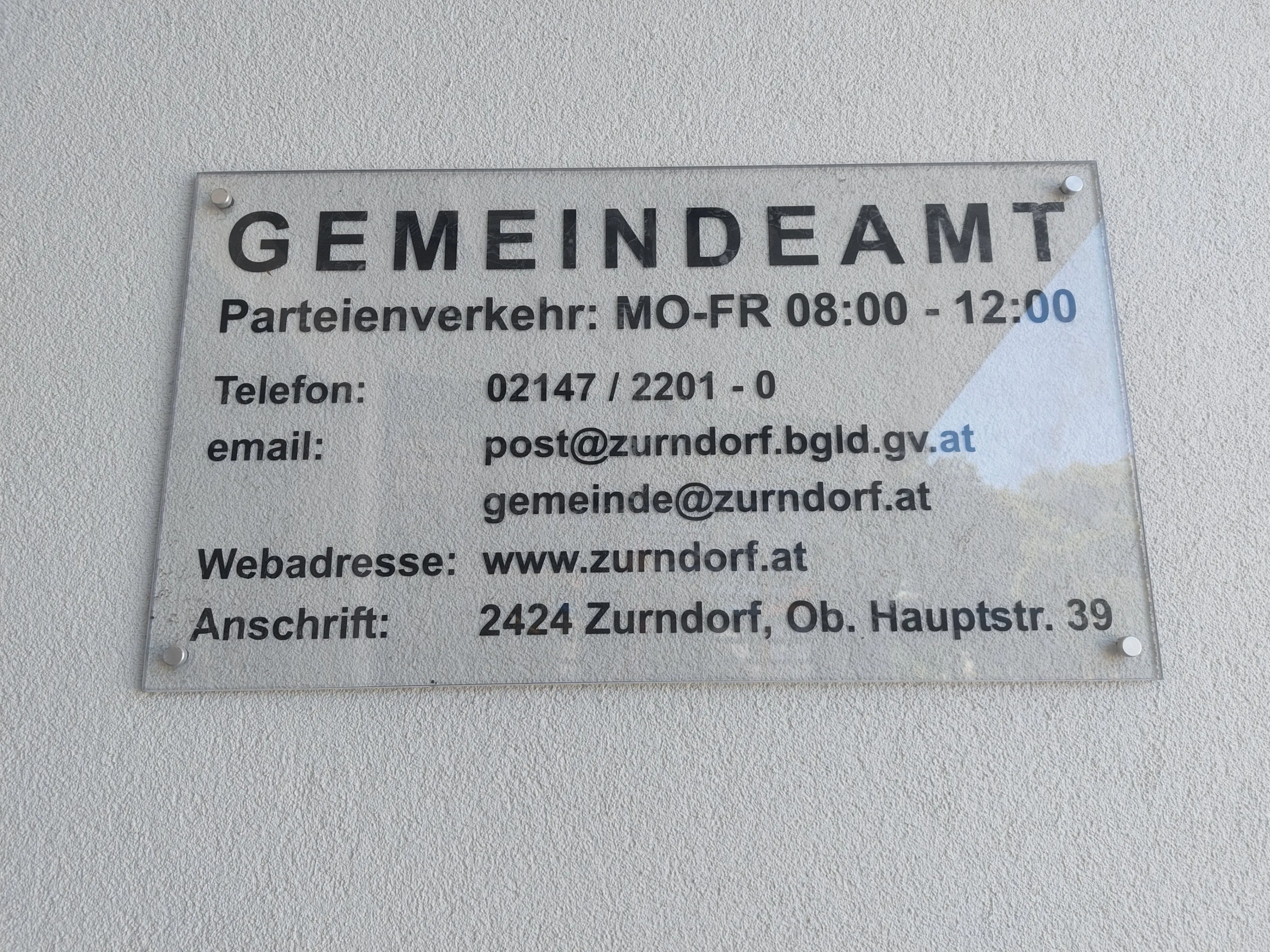 Schild für Gemeindeamt mit Kontaktdaten. Geöffnet Mo-Fr von 08:00 bis 12:00. Telefonnummer 02147/2201-0. E-Mails: post@zurndorf.bgld.gv.at und gemeinde@zurndorf.at. Webseite: www.zurndorf.at. Adresse: 2424 Zurndorf, Obere Hauptstr. 39.