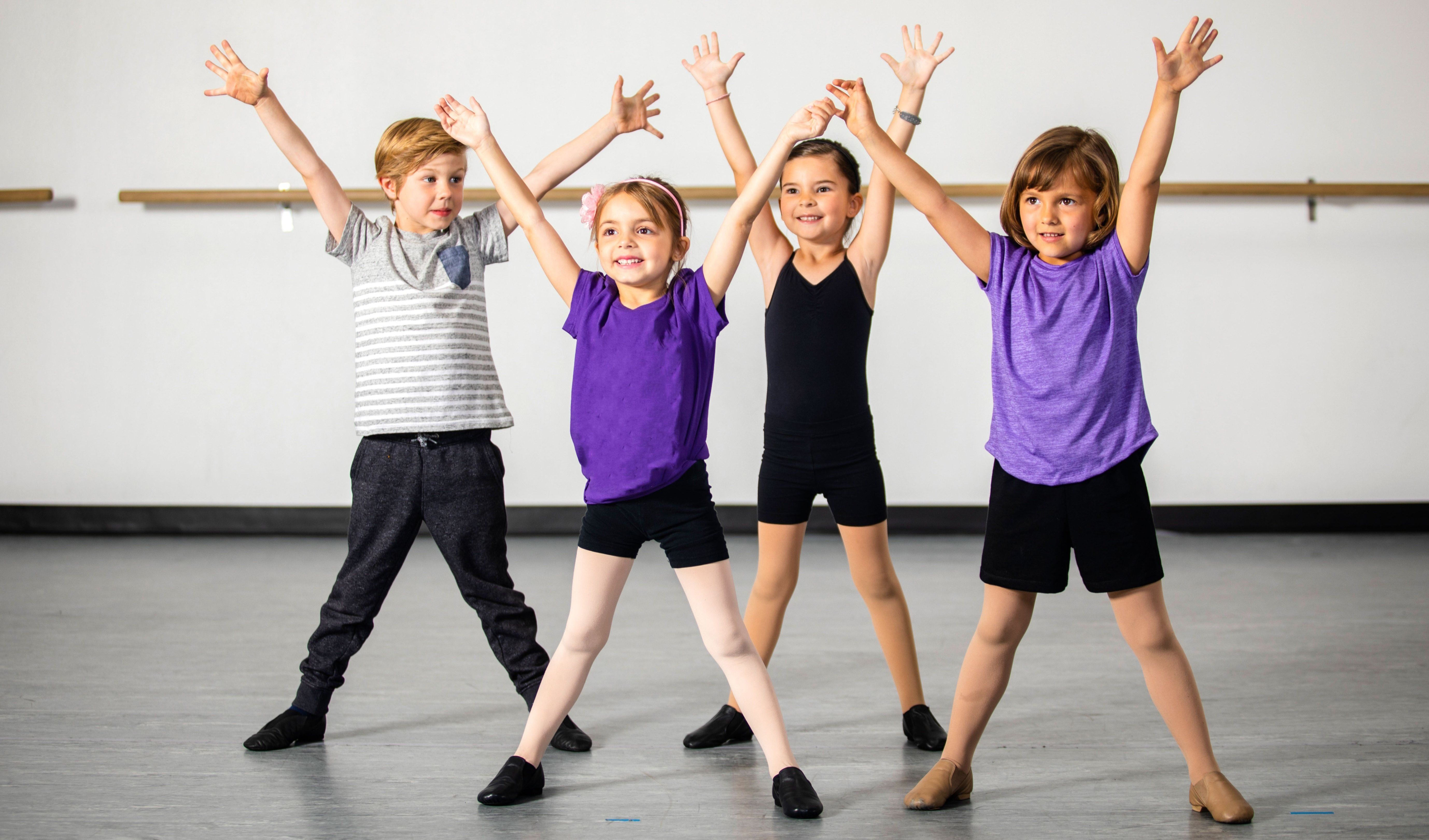 Vier Kinder in einem Tanzstudio, zwei Mädchen in lila und schwarzen Shirts, eines in einem schwarzen Shirt und ein Junge in einem grauen Shirt, alle mit erhobenen Händen.