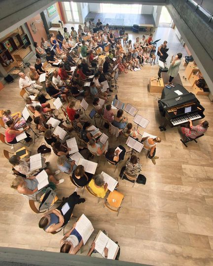 Eine große Gruppe von Menschen ist in einem Raum versammelt, einige sitzen und andere stehen, alle halten Musikpartituren. Ein Pianist spielt an einem Klavier, und ein Dirigent steht in der Nähe.