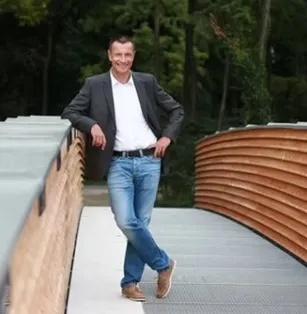 Ein Mann im Anzug steht auf einer Brücke, lächelt und posiert für ein Foto.