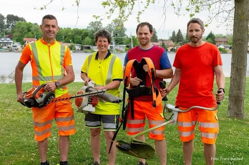 Vier Personen in Warnkleidung stehen in einem Park und halten verschiedene Gartenwerkzeuge in der Hand. Der Mann auf der linken Seite hält eine Kettensäge, während die anderen Trimmer und einen Rucksack halten. Sie lächeln und scheinen für ein Foto zu posieren.