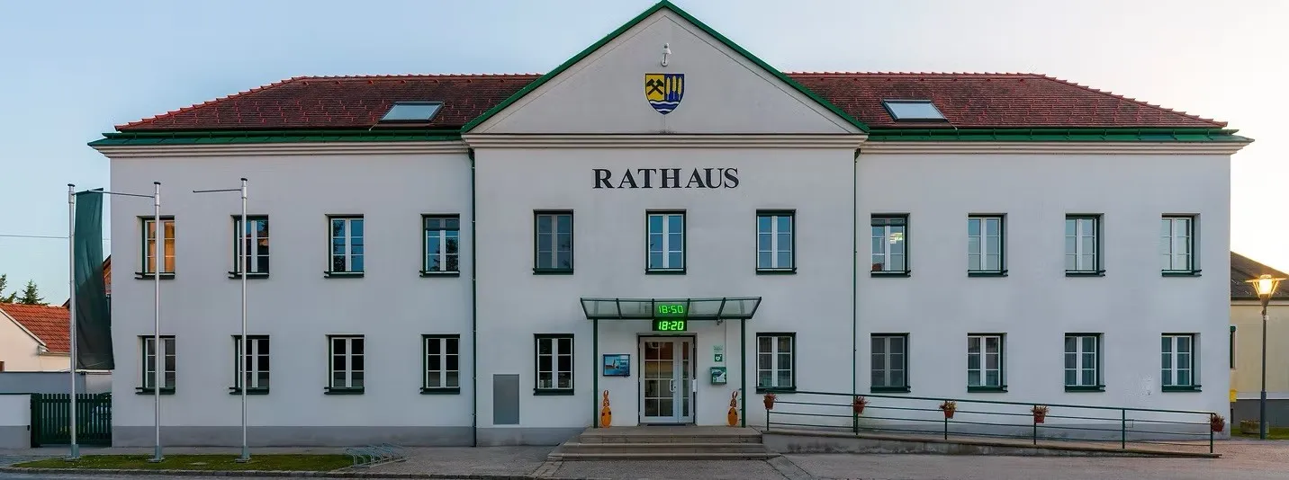 Die Vorderseite eines weißen Gebäudes mit dem Wort Rathaus darauf. Es hat ein rotes Dach, grüne Verzierungen und mehrere Fenster. Das Gebäude hat eine Glastür mit einer digitalen Uhr darüber.