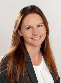 Eine Frau mit langen braunen Haaren, die ein weißes Blusenhemd und einen grauen Blazer trägt, lächelt und schaut nach vorne.