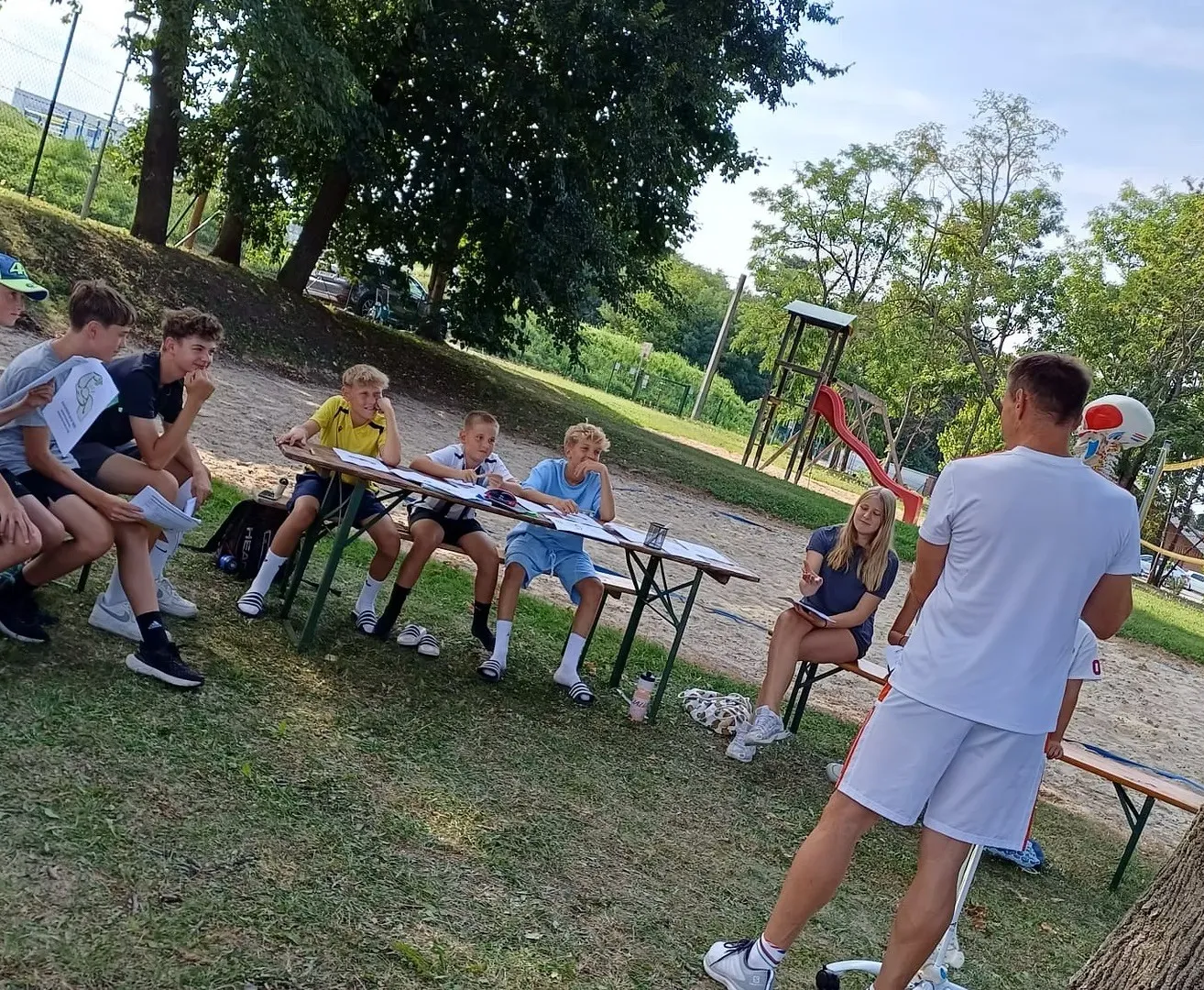 Eine Gruppe von Kindern sitzt um einen Tisch in einem Park, ein Mann steht in der Nähe. Sie scheinen eine Art Aktivität oder Test zu machen.