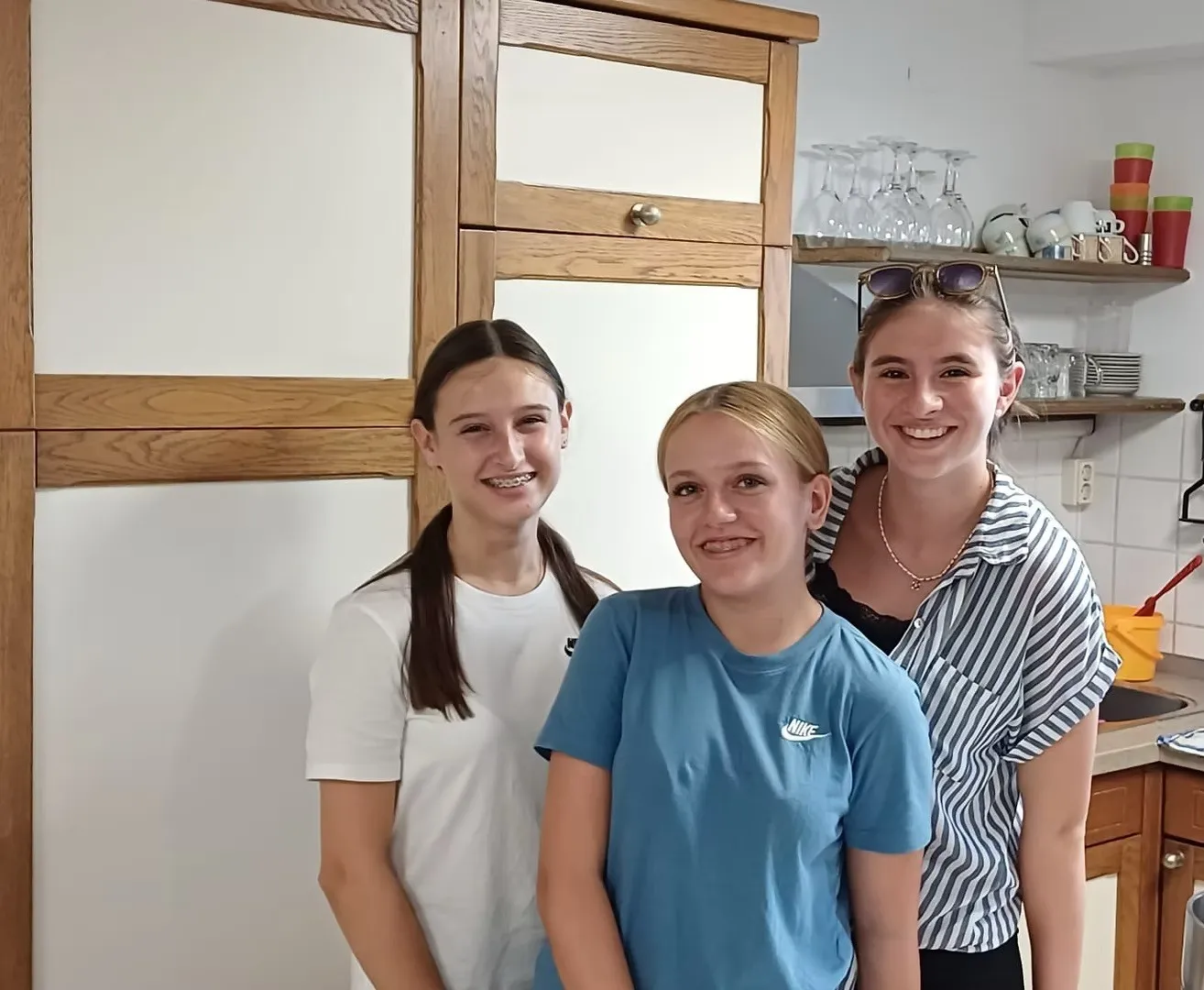 Drei junge Frauen stehen gemeinsam in einer Küche und lächeln für ein Foto. Die Frau auf der linken Seite trägt ein weißes T-Shirt, die mittlere ein blaues Nike-Shirt und die rechte ein gestreiftes Shirt. Hinter ihnen sind Holzschränke und Regale mit Gläsern zu sehen.