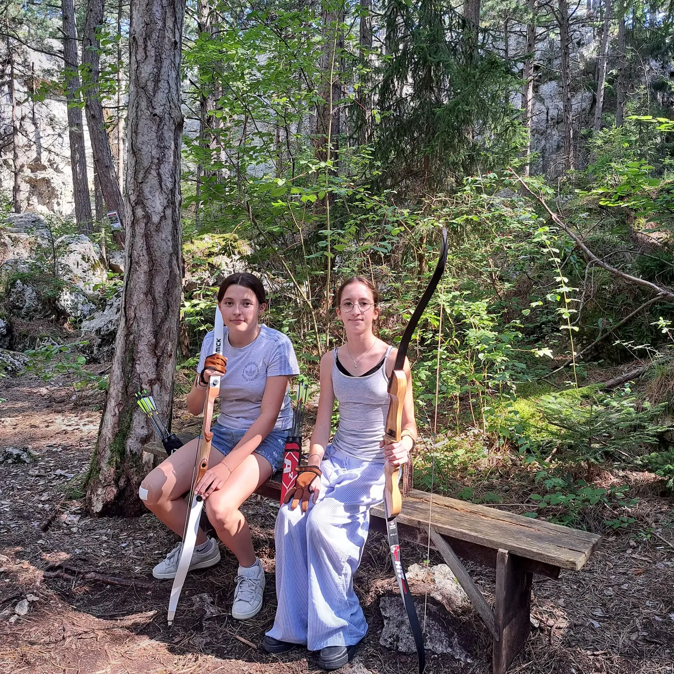 Zwei Frauen sitzen auf einer Holzbank im Wald, beide halten Bögen und Pfeile. Eine trägt ein graues Shirt und Jeansshorts, die andere ein ärmelloses Top und Hosen. Sie schauen in die Kamera.