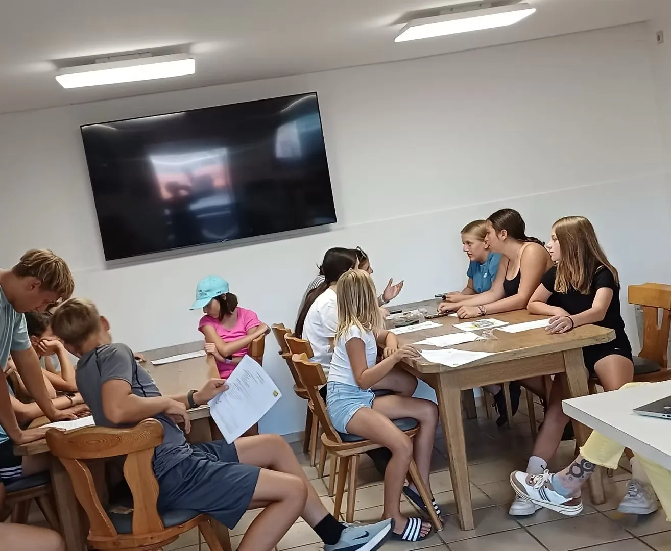 Eine Gruppe junger Mädchen und Jungen sitzt um einen Tisch in einem Klassenzimmer und diskutiert möglicherweise etwas. Ein Monitor ist über ihnen an der Wand montiert.