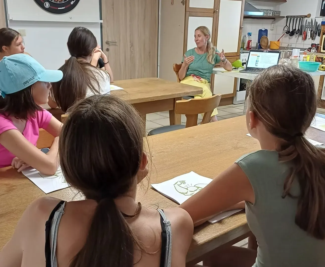 Eine Frau unterrichtet eine Gruppe junger Mädchen in einer Küche. Die Mädchen sitzen an einem Tisch und hören aufmerksam zu. Die Lehrerin gestikuliert mit den Händen, während sie spricht.