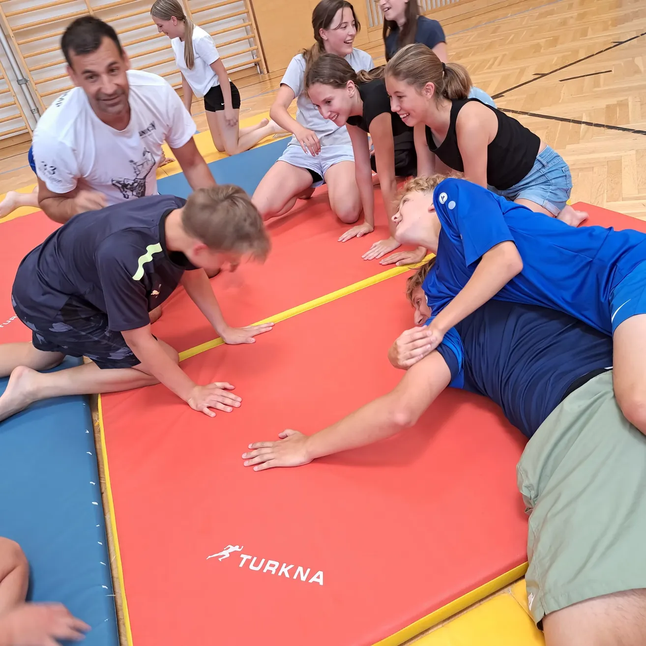 Eine Gruppe von Kindern und Erwachsenen übt Gymnastik auf einem roten und blauen Teppich. Sie lächeln und scheinen Spaß zu haben.