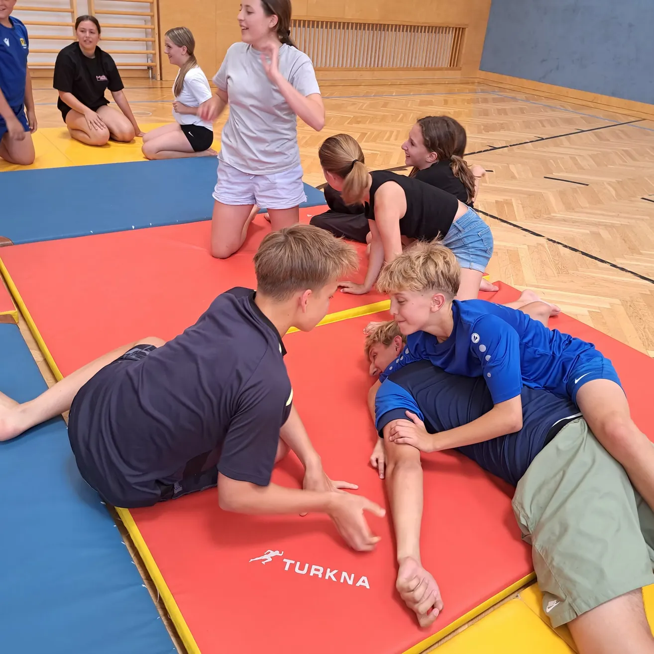 Eine Gruppe junger Menschen praktiziert Yoga auf Matten in einem Fitnessstudio. Ein Junge liegt auf dem Rücken, während ein anderer Junge kniet und zeigt.