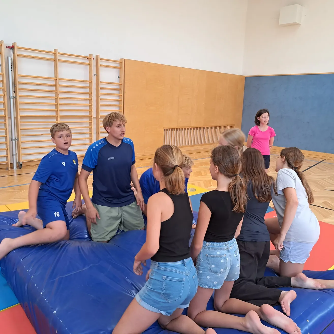 Eine Gruppe von Kindern und Jugendlichen versammelt sich auf einer blauen Luftmatratze in einer Turnhalle, wobei zwei Jungen in blauen Trikots sitzen und andere stehen. Ein Mädchen in einem rosa Shirt steht in der Nähe.