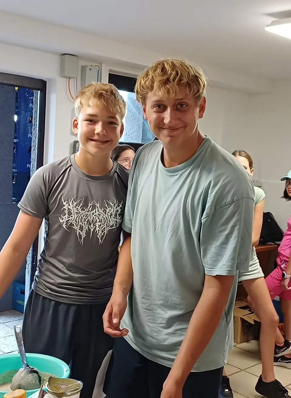 Zwei junge Jungen stehen eng beieinander und lächeln für ein Foto. Einer trägt ein graues T-Shirt mit einem weißen Design, der andere ein hellblaues T-Shirt. Hinter ihnen sind eine Frau und ein Mädchen teilweise zu sehen.
