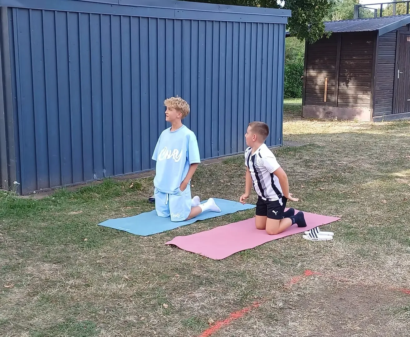 Zwei Jungen knien auf Yogamatten in einem Außenbereich, einer trägt ein hellblaues T-Shirt mit weißem Text und der andere ein schwarz-weiß gestreiftes T-Shirt.
