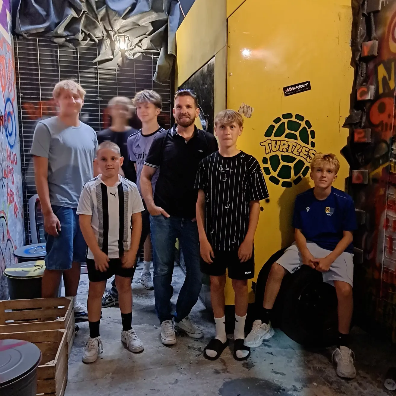 Eine Gruppe junger Jungen und Männer posiert für ein Foto vor einer gelben Wand mit Graffiti und einem Schildkröten-Logo.