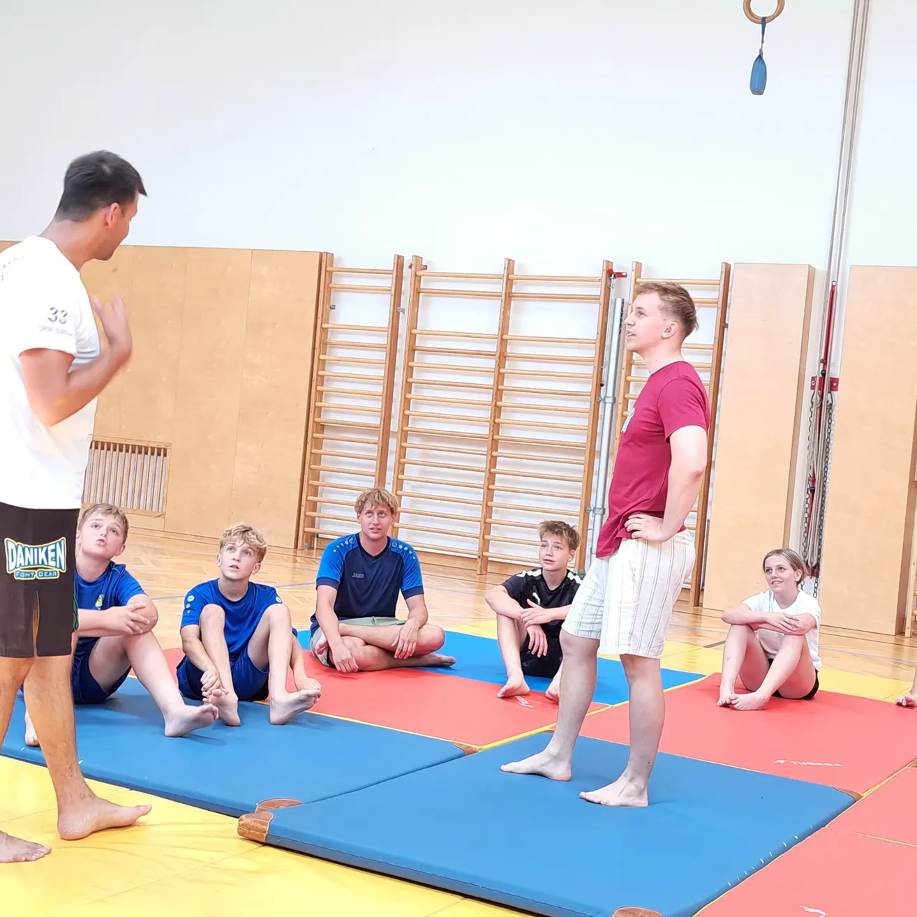 Eine Gruppe junger Männer und Frauen sitzt auf Matten in einem Fitnessstudio und hört zwei Männern zu, die vor ihnen stehen.
