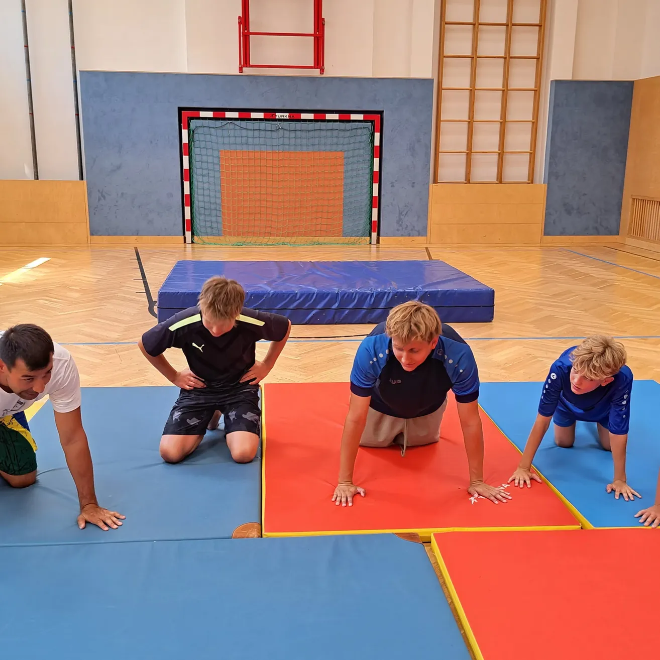 Vier junge Männer machen Liegestütze auf farbigen Matten in einer Turnhalle. Einer trägt ein schwarzes Shirt, ein anderer ein blaues und zwei tragen weiße. Ein Tor steht im Hintergrund.