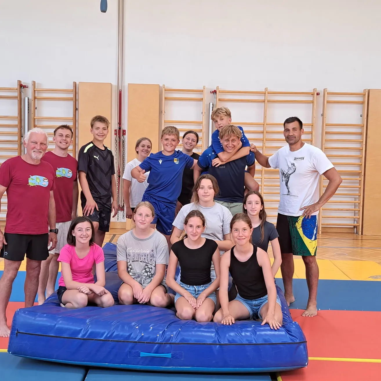 Eine Gruppe junger Menschen und Erwachsener posiert für ein Foto in einem Fitnessstudio, wobei einige auf einer blauen Matte sitzen und andere dahinter stehen.
