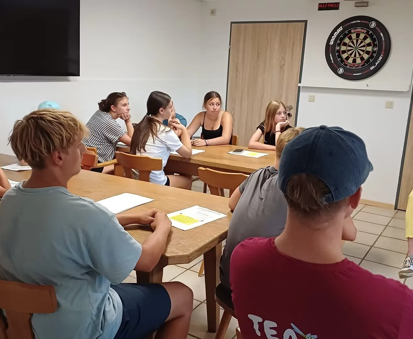 Eine Gruppe junger Menschen sitzt um einen Tisch in einem Raum, wahrscheinlich in einer Besprechung. Papiere und ein Dartboard sind sichtbar.