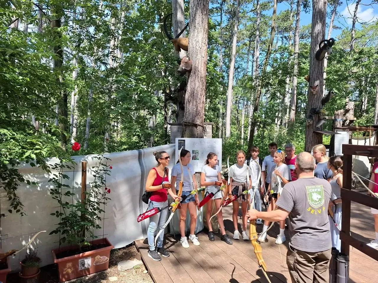 Eine Gruppe junger Menschen und ein erwachsener Mann üben Bogenschießen in einem Wald. Sie stehen auf einer Holzplattform mit einem weißen Hintergrund.