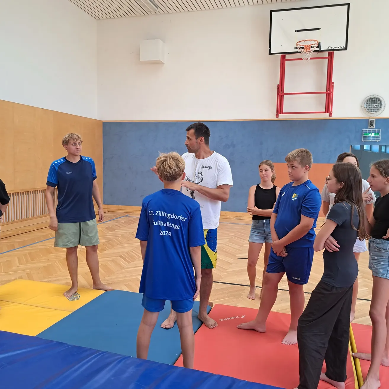 Eine Gruppe junger Menschen und ein Mann in einer Turnhalle, der Mann spricht zu ihnen, während sie auf Matten stehen.