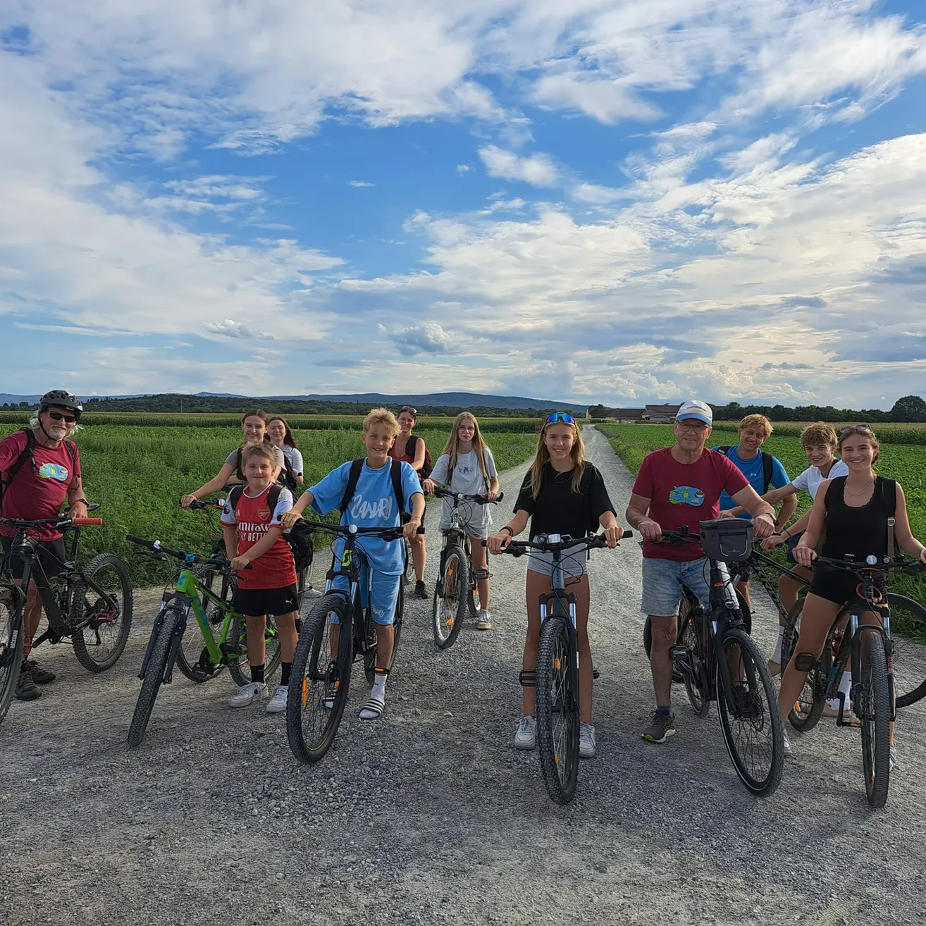 Eine Gruppe von Menschen, einschließlich Kindern, steht mit ihren Fahrrädern auf einem Kiesweg in einem Grasfeld unter einem blauen Himmel mit Wolken.