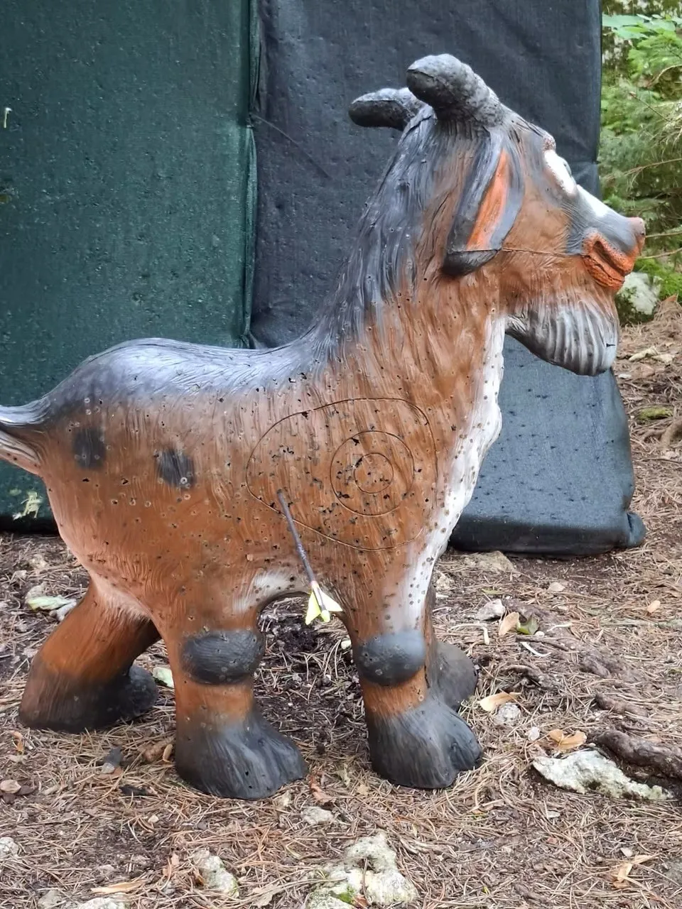 Eine lebensgroße Ziegenstatue mit Einschusslöchern und einem Pfeil im Oberkörper, die auf einem Erdboden steht.